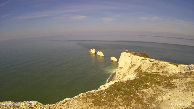 A panoramic view of white chalk cliffs by the ocean, with green patches on the top of the cliffs and a small building on the right. In the distance, there is a lighthouse on a set of rocks protruding from the water, and the sea appears calm with gentle waves. The sky is mostly clear with some wispy clouds. The image has a timestamp at the bottom left corner, and the website "isleofight.com" is displayed at the bottom right.