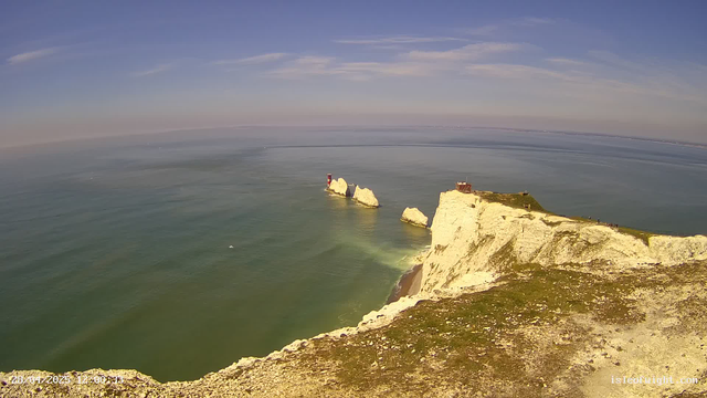 A panoramic view of white chalk cliffs along the coastline, extending into the sea. Several large rocks and sea stacks are visible offshore, with one featuring a small lighthouse or beacon. A small building is situated at the top of the highest cliff. The sky above is mostly clear with some wispy clouds, and the sea is calm with gentle waves.