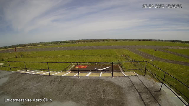 An image from a webcam showing the view of a grassy airfield with a clear sky and some clouds. In the foreground, there is a rooftop with a railing, and below it, a small helicopter landing pad with a red mark. The airfield extends into the distance with a paved runway and green fields surrounding it. The top right corner displays the date and time as "28-04-2025 12:00:35". The bottom left corner has the text "Leicestershire Aero Club".
