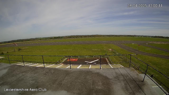 A wide view of an airfield with a paved runway extending into the distance. In the foreground, there is a parking area and a small helipad with a white helicopter parked on it. A metal railing surrounds the helipad. To the left, a flag is blowing in the wind. The sky is partly cloudy with patches of blue. The image includes a timestamp showing "28-04-2025 13:00:11" in the top right corner and the text "Leicestershire Aero Club" in the bottom left corner.