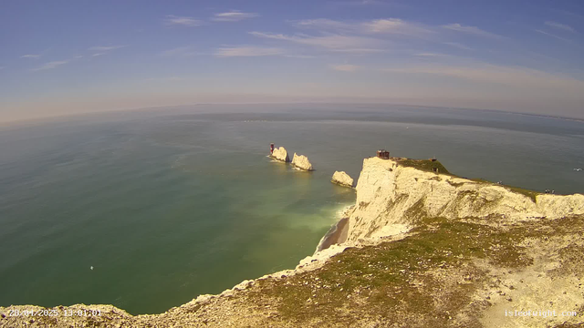 A view of white chalk cliffs extending into the sea under a partly cloudy sky, with a lighthouse and a small building atop the cliffs. The ocean is calm and greenish, with a distant horizon.  
The image appears to be taken from a webcam, with a timestamp and website watermark at the bottom.