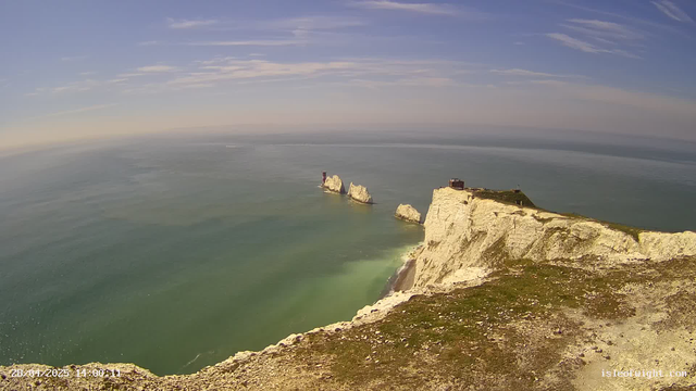 Aerial view of white chalk cliffs along the coastline, with a narrow grassy area and structures on top. The cliffs extend into the sea, which is calm and greenish in color. In the distance, large white rock formations emerge from the water. The sky is mostly clear with some wispy clouds, and a faint horizon line is visible where the sky meets the sea.