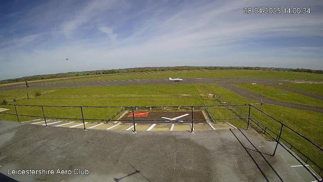 A view of an airfield with a small airplane taxiing on the runway in the distance under a partly cloudy sky. The image is taken from a webcam at the Leicester Shire Aero Club, with a timestamp of April 28, 2025, at 14:00:34. In the foreground, there is a ramp area with parking spaces and safety barriers.