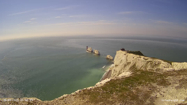 An aerial view of white chalk cliffs extending into the sea, with a small building or structure on top of the cliffs. Several large rock formations are visible offshore in the water. The sky is mostly clear with a few wispy clouds, and the calm sea reflects sunlight. The scene is captured during daytime.