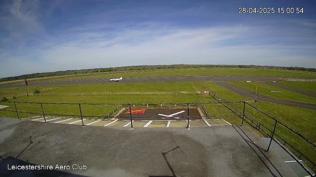 An aerial view of a small airport with a single white airplane on the runway. The sky is mostly clear with some wispy clouds. In the foreground, there is a parking area with several marked spaces and a fenced-off section. The location is identified as Leicestershire Aero Club, and the timestamp shows the date as April 28, 2025, at 3:00 PM.