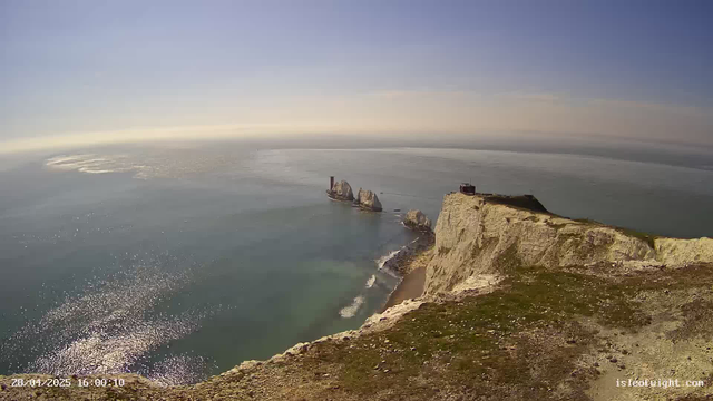 A view from the top of white chalk cliffs overlooking the ocean. The cliffs are grassy and steep, with a small structure near the edge. In the distance, there are notable rock formations rising from the water, one with a chimney-like structure. The sky is clear with a subtle gradient from blue to pale yellow near the horizon. Sunlight reflects off the water, creating a shimmering effect.
