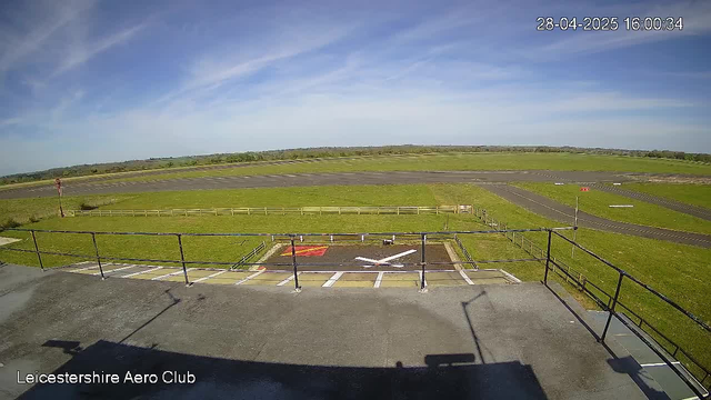 An outdoor view of a grass airfield under a partly cloudy blue sky. In the foreground, there is a small paved area with a white arrow painted on the ground pointing to the right and a red area with a white "X" mark. Surrounding this paved area is a metal safety railing. The background shows open green fields and a runway extending into the distance. There is a timestamp in the top right corner reading "28-04-2025 16:00:34," and text in the bottom left corner that says "Leicestershire Aero Club."
