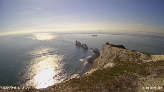 A scenic view of a coastal landscape with white chalk cliffs and a grassy area at the top. The ocean stretches out into the horizon, reflecting sunlight, with a few boats visible in the distance. The sky is mostly clear with some light clouds, and the overall scene is bathed in bright sunlight. The image includes a timestamp of "28/04/2025 17:00:24" and a website watermark "isleofwight.com" at the bottom right corner.