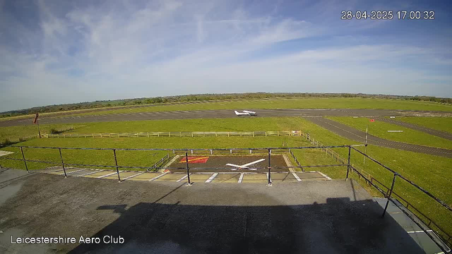 A wide view of an airport runway and grassy area under a partly cloudy sky. In the foreground, a section of an observation deck with railings and a marked landing zone. An airplane on the runway in the distance, with another airplane taxiing or parked further back. The image is timestamped "28-04-2025 17:00:32" and labeled "Leicestershire Aero Club."