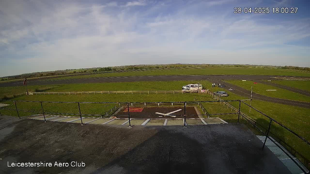 A view from a webcam overlooking an outdoor area at Leicestershire Aero Club on a clear day. The sky is partly cloudy with some wispy clouds. In the foreground, there is a small helipad with a white helicopter parked on it. Beyond the helipad, there are grassy fields with a few parked cars, a small pile of equipment, and a walking path. The horizon shows open fields and a distant line of trees, with rolling hills in the far background. The timestamp on the image indicates it was taken on April 28, 2025, at 6:00 PM.