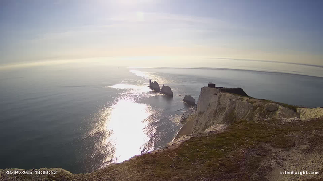 A view of towering white chalk cliffs over the ocean, with the sun reflecting off the water's surface. There are several large sea stacks and rock formations near the shoreline, and a small structure is visible on top of the cliffs. The sky is clear with some light clouds, and the overall scene captures a serene coastal landscape during seasunny weather.