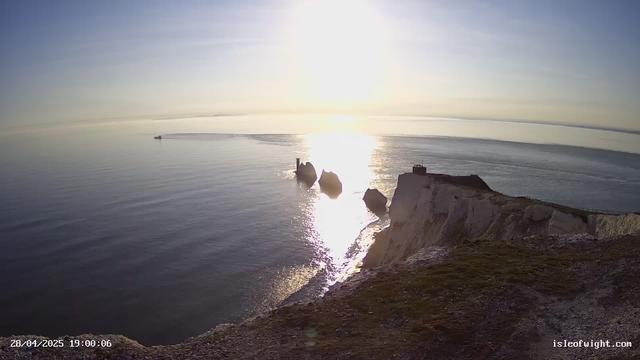 A view of the ocean at sunset or sunrise, with the sun low on the horizon casting a bright reflection on the water. There are white chalk cliffs along the shoreline, with a small building or structure on top of one of the cliffs. A helicopter is flying near the cliffs, and a boat is visible in the distance on the water. The image has a timestamp in the bottom left corner and a website URL in the bottom right corner.