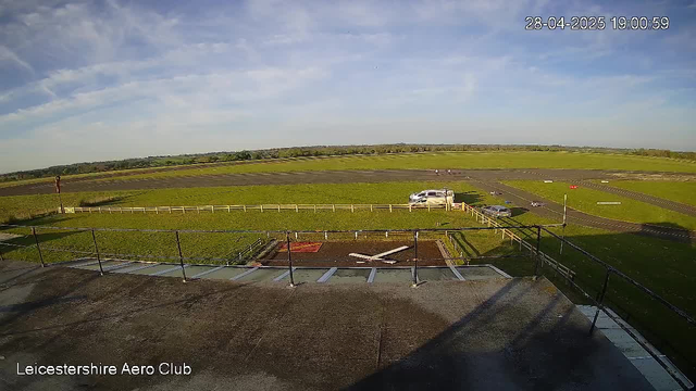 An outdoor view from a webcam showing a grassy airfield with a few parked white and black vehicles, a small fenced area, and a distant horizon with green fields under a partly cloudy sky. The timestamp in the top right corner reads "28-04-2025 19:00:59," and the bottom left corner has the text "Leicestershire Aero Club."
