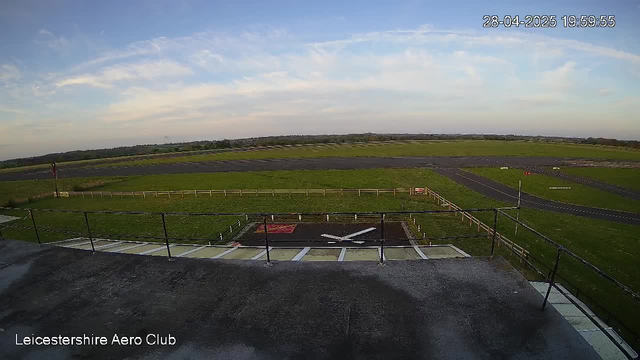 A wide-angle view of an open outdoor area shows a clear sky with some scattered clouds. In the foreground, there is a raised platform with a black railing and a small landing pad that has markings resembling a helicopter landing zone. The landing zone is painted black with white outlines and a large "H" in white. The platform appears to be part of Leicester Aero Club, as indicated by the text in the bottom left corner. In the distance, there is a grassy field enclosed by a fence, with a few small signs and markers. Beyond the field, a road curves through the landscape, which extends toward the horizon with some gentle rolling hills. The top right corner displays a timestamp: "28-04-2025 19:59:55."