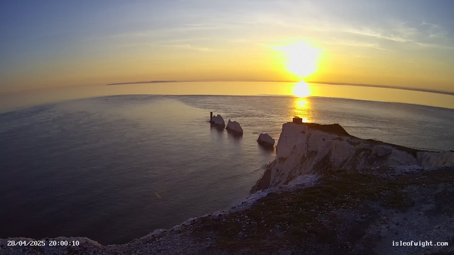 A serene scene of white chalk cliffs overlooking the ocean at sunset. The sun is low on the horizon, casting a golden glow across the sky and reflecting on the calm water below. There are several large rocks emerging from the sea near the base of the cliffs. The timestamp in the lower left corner reads "28/04/2025 20:00:10," and the website "isleofwight.com" is displayed in the lower right corner.
