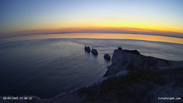 A panoramic view of the sea at sunset with a series of white chalk cliffs in the foreground. Several large rocks are visible in the water near the cliffs, and a small lighthouse with a red light stands on a rock formation. The sky displays a gradient of colors from deep blue to orange and yellow near the horizon.