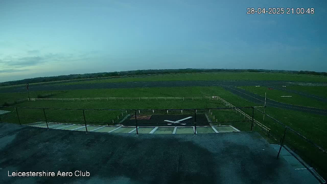 A view from a webcam showing the Leicester aerodrome at dusk with a small runway and grassy fields extending into the distance. The sky is mostly clear with some clouds, and the timestamp indicates it is 9:00 PM on April 28, 2025. There is a small helipad or landing pad in the foreground with parking spaces nearby. The bottom left corner displays the text "Leicestershire Aero Club."