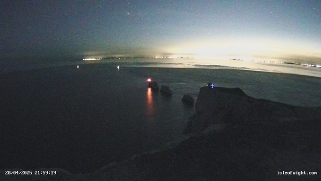 A nighttime scene of a calm ocean with large rocks in the foreground. The horizon shows a faint glow from the setting or rising sun, with distant city lights on the shoreline. Several small navigation beacons with red and blue lights are visible on the rocks, casting reflections on the water. The sky is clear with faint stars visible. The timestamp in the bottom left corner reads "28/04/2025 21:59:39", and the website "isleofwight.com" is displayed in the bottom right corner.