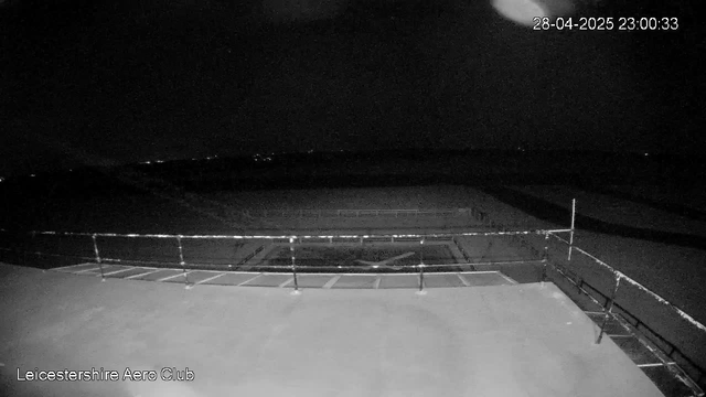 A black-and-white image taken at night shows a fenced-off area with a flight or sports field in the distance. The scene appears dark, with very limited lighting, and the sky is mostly black with the moon partially visible in the top right corner. The date and time stamp in the top right corner indicates it was taken on April 28, 2025, at 23:00:33. The lower left corner has the text "Leicestershire Aero Club".