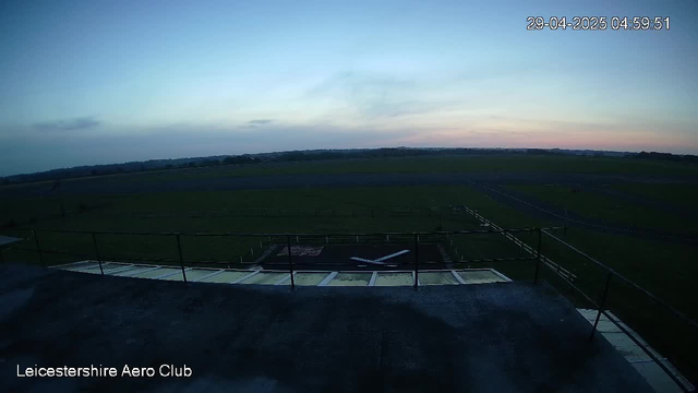 A wide view of an open grassy airfield during twilight with a small aircraft on a runway. The sky is a gradient of soft blue and pink hues, and there are a few scattered clouds. A railing is visible in the foreground, and the text "Leicestershire Aero Club" is at the bottom left corner. The timestamp in the upper right corner reads "29-04-2025 04:59:51."
