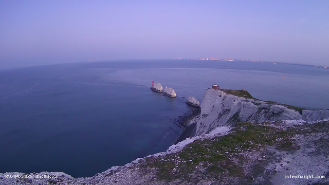 A view of white chalk cliffs and rocks extending into the ocean at dawn or dusk, with a lighthouse on one of the rocks and a small building near the edge of the cliffs. The sky is pale, and the water is calm with a few small boats visible in the distance. The scene has a soft, tranquil atmosphere with subtle lighting.
