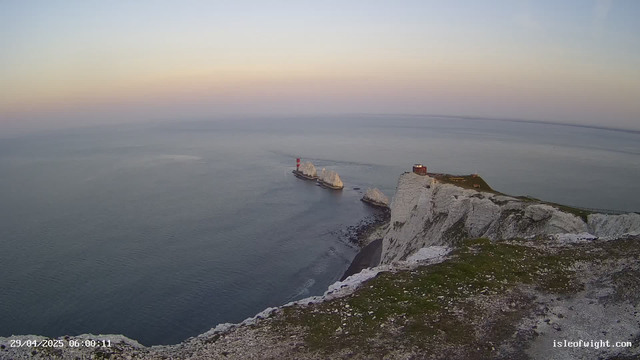 A panoramic view of white chalk cliffs along the coastline, with a small building on top and a pathway. There are three large rock formations extending into the calm sea, one with a red and white striped lighthouse on it. The sky is clear, and the scene appears to be during sunrise or sunset, with soft lighting. The timestamp on the image reads "29/04/2025 06:00:11," and the website "isleofwight.com" is visible in the bottom right corner.