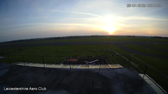 A sunrise over an open grassy field with a small helipad and a propeller aircraft parked nearby. The scene is viewed from an elevated platform with railings. The sky has a few wispy clouds and the timestamp indicates it is early morning. The words "Leicestershire Aero Club" are visible in the bottom left corner.