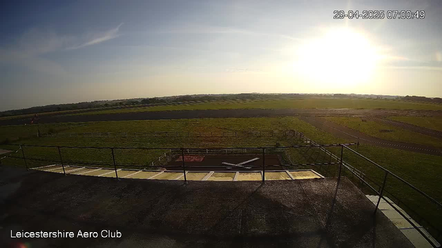 An outdoor view of a flat grassy field under a bright, low sun in the sky. Some small paved pathways or runways are visible spanning across the field. There is a black metal railing along the edge of a concrete platform in the foreground. The image is timestamped "29-04-2025 07:00:49" in the upper right corner, and "Leicestershire Aero Club" is written in the bottom left corner.