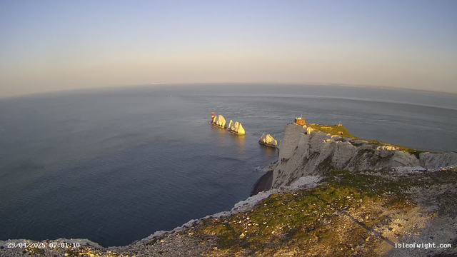 A wide view of white chalk cliffs with grassy tops extending over the ocean, with large white rock formations jutting out into the water. The scene is bathed in warm sunlight, suggesting early morning or late afternoon. The sky is clear with no clouds, and the water below is calm, reflecting the sky. There is a small structure on the edge of the cliff, and the date and time are displayed at the bottom left corner.