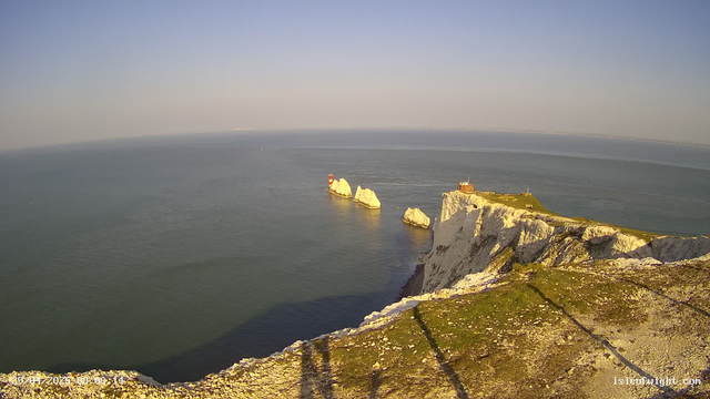 A high-angle view of white chalk cliffs along a coastline, with a grassy top and a small building near the edge. Several large white rocks are visible in the water off the coast, and a red and white lighthouse stands on a rock formation in the sea. The sky is clear and the water is calm.