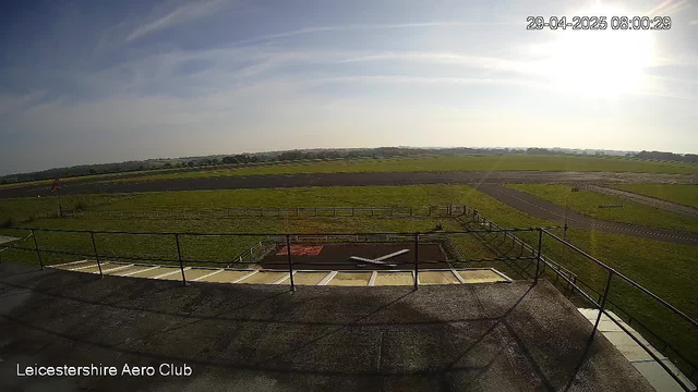 An outdoor view from a webcam shows a bright, sunny day at Leicesterhire Aero Club. The scene includes a paved area with a metal railing, a small hatch in the ground, and a grassy airfield with runways and taxiways extending into the distance. The sky is mostly clear with some wispy clouds. The timestamp on the image indicates the date and time as April 29, 2025, at 8:00 AM.
