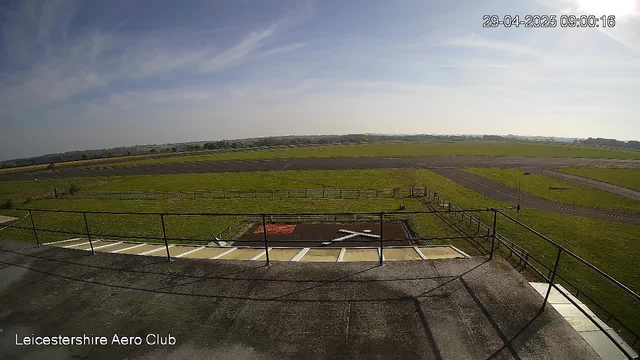 An empty outdoor airfield or runway surrounded by grassy fields, with a small fenced area and a ramp leading down from a concrete platform. The sky is mostly clear with some wispy clouds. The top right corner shows a timestamp of "29-04-2025 09:00:16," and the bottom left corner indicates "Leicestershire Aero Club."