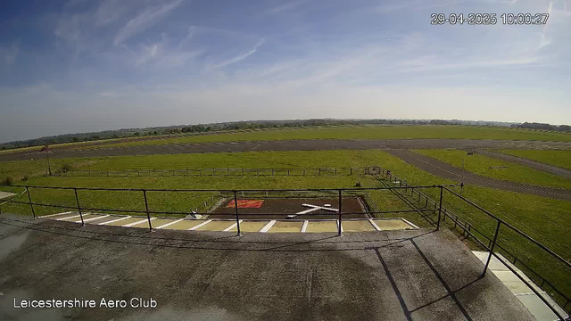 A wide view of an airfield with grassy runways and taxiways stretching into the distance under a partly cloudy sky. In the foreground, there's a rooftop with a railing, some painted lines, and a small helipad area marked with a large white "H". The image includes a timestamp in the upper right corner showing "29-04-2025 10:00:27", and the bottom left corner has the text "Leicestershire Aero Club".