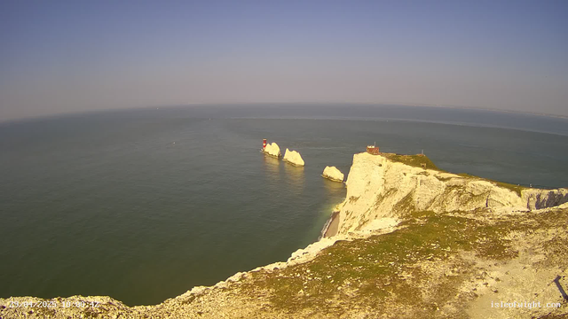A panoramic view of white chalk cliffs along a coastline, with a small lighthouse and buildings at the edge. The cliffs jut out into the calm sea, which is dotted with a few small boats. The sky is clear with a subtle gradient from blue to a faint haze near the horizon.