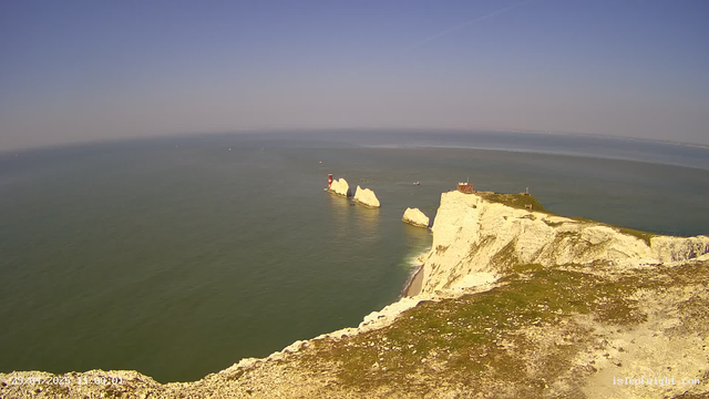 A scenic view of white chalk cliffs along the coastline, with several large white rock formations extending into the greenish-blue sea. A small building and a lighthouse are situated on the cliff edge. The sky is clear with no clouds.