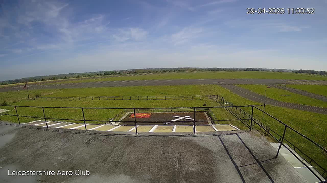 An outdoor view of a grassy airfield with a runway extending into the distance under a partly cloudy sky. In the foreground, there is a parking area with parking spaces and a small helipad marked with a white helicopter symbol. A black railing runs along the edge of the parking area. The image is timestamped "29-04-2025 11:00:52" in the top right corner, and the text "Leicestershire Aero Club" appears at the bottom left corner.