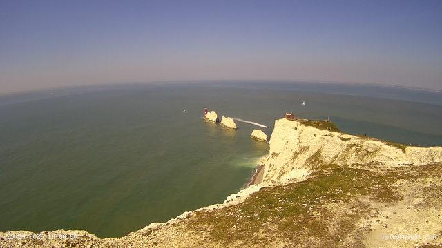 A wide view of white chalk cliffs extending into the sea with greenish water below. There is a lighthouse on a small rock formation near the cliffs, and a building on top of the cliffs. Several sailboats are visible in the distance under a clear blue sky. A boat is leaving a white wake behind it, heading away from the cliffs.