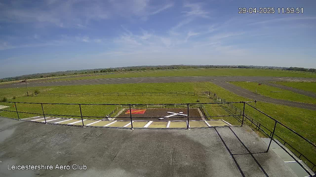 A wide view of an open grassy airfield under a partly cloudy blue sky, with a small helicopter landing pad marked with a white "H" and red nearby signage in the foreground. There is a paved area with a railing, and distant fields and a horizon line in the background. The image is timestamped "29-04-2025 11:59:51" and labeled "Leicestershire Aero Club" in the bottom left corner.