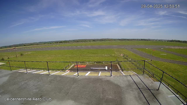 An outdoor view from a webcam at Leicestershire Aero Club shows a grassy airfield with a paved runway stretching across the scene under a partly cloudy blue sky. There is a small parking area with white painted lines and a safety railing in the foreground. The timestamp in the top right corner reads "29-04-2025 12:59:51."