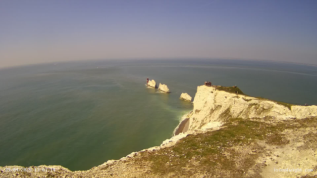 Aerial view of white chalk cliffs along the coastline with grassy tops, extending into the ocean. There are several large rock formations protruding from the water near the cliffs, including a small lighthouse on one of the rocks. The sea is calm with a greenish tint, and the sky is clear with a soft blue haze near the horizon.