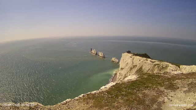 Aerial view of white chalk cliffs with a narrow pathway on top, overlooking the ocean. The cliffs are steep and rugged, with some patches of greenery on the top. In the distance, white chalk stacks stand in the water off the coast, surrounded by calm, shimmering sea under a clear blue sky.
