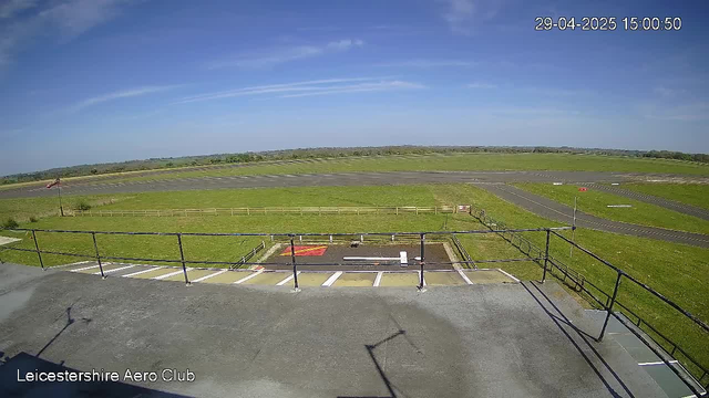 An expansive view of a clear day at an airfield shows an open grassy area and a paved race track in the distance. The sky is mostly blue with some wispy clouds. In the foreground, there is a small parking area with marked parking spaces and a fenced section. The timestamp indicates it is April 29, 2025, at 3:00 PM, and the location is the Leicestershire Aero Club.