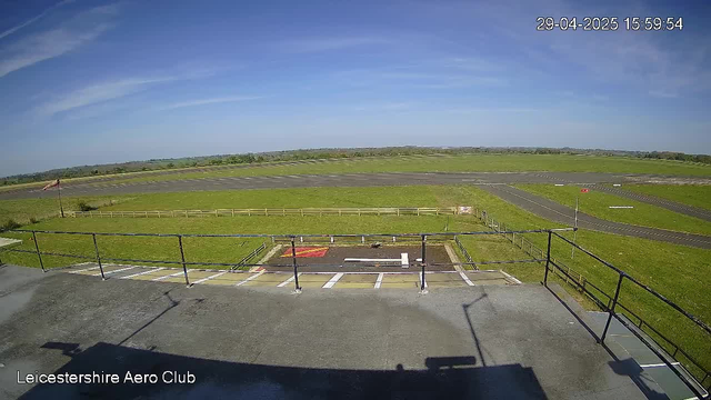 An outdoor view from a webcam at Leicesterhire Aero Club showing a clear blue sky with some wispy clouds, a grassy airfield, runways, and taxiways stretching into the distance. There is a small paved area with parking spaces and a railing in the foreground. The timestamp in the upper right corner indicates the date and time as April 29, 2025, at 3:59 PM.
