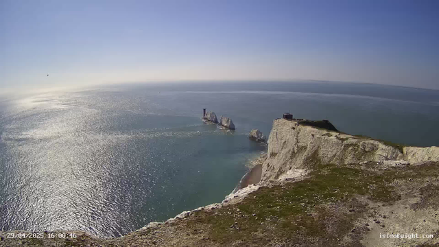 A panoramic view of white chalk cliffs along the coastline, with a small building on the edge. The cliffs descend to a beach, and the ocean extends into the horizon with sunlight reflecting off the water. There are several sea stacks rising from the sea near the cliffs, and the sky is clear and blue.