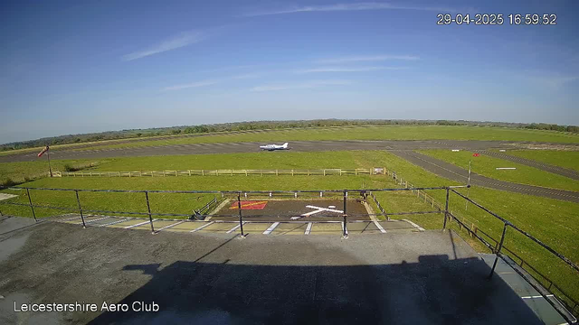 An airplane taxiing on the runway at Leicestershire Aero Club. The sky is mostly clear with some wispy clouds, and the scene is sunny. The photo is taken from an elevated position with a black railing at the bottom. There are parking spaces with painted lines in the foreground, and a windsock on a pole and runway markings are visible. The timestamp in the top right corner reads "29-04-2025 16:59:52."
