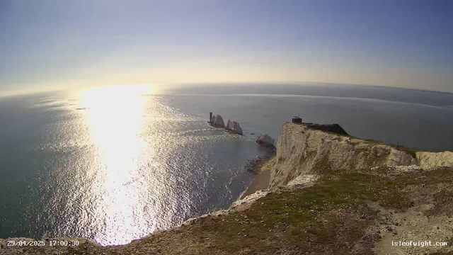 Aerial view of white chalk cliffs along the coastline at sunset, with the sun reflecting brightly on the ocean. The cliffs extend along the shoreline, with a small building and a tower visible on top of one of the cliffs. The ocean stretches out to the horizon, with rocks and sea stacks near the base of the cliffs. The sky is clear and the scene is bathed in warm, golden light.