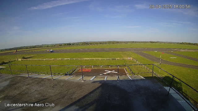 A view from a webcam showing an airfield with a grassy runway, a small airplane in the distance, and a clear blue sky with some wispy clouds. The image is timestamped "29-04-2025 18:00:56" and is labeled "Leicestershire Aero Club" at the bottom left corner.