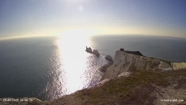 A panoramic view of white chalk cliffs overlooking the ocean during sunset. The sun is low on the horizon, reflecting bright light on the water. Some small rock formations and a lighthouse are visible on the cliffs. The sky is clear with a faint hint of clouds, and the overall scene exudes a calm, serene atmosphere.