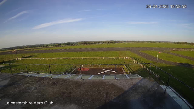 A view from a weather surveillance camera at Leicestershire Aero Club showing a clear sky with a few wispy clouds. In the foreground, there is a small section of a rooftop with safety railings. Beyond the rooftop, there is a green grassy area with a marked section, possibly a helipad or small landing area, enclosed by a white fence. In the background, there is a runway with a red and white windsock on a pole and several small advertisements or signs along the edge. The image is timestamped April 29, 2025, at 6:59:58 PM.