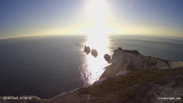 A wide view of the ocean at sunset, with bright sunlight reflecting off the water. White chalk cliffs are visible in the foreground, with a small building or structure on the edge of the cliffs. Several large rocks are present in the water near the cliffs. The sky is mostly clear with a gradient from blue to warm hues near the horizon. The image includes timestamp information in the bottom left corner and a website URL in the bottom right corner.